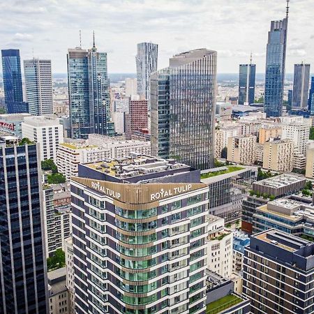 Royal Tulip Warsaw Centre 아파트 호텔 외부 사진 The photo shows a panoramic view of a modern city skyline. In the foreground, there are several tall buildings with contemporary architectural designs. One building prominently features the name "Royal Tulip." The background is filled with numerous s
