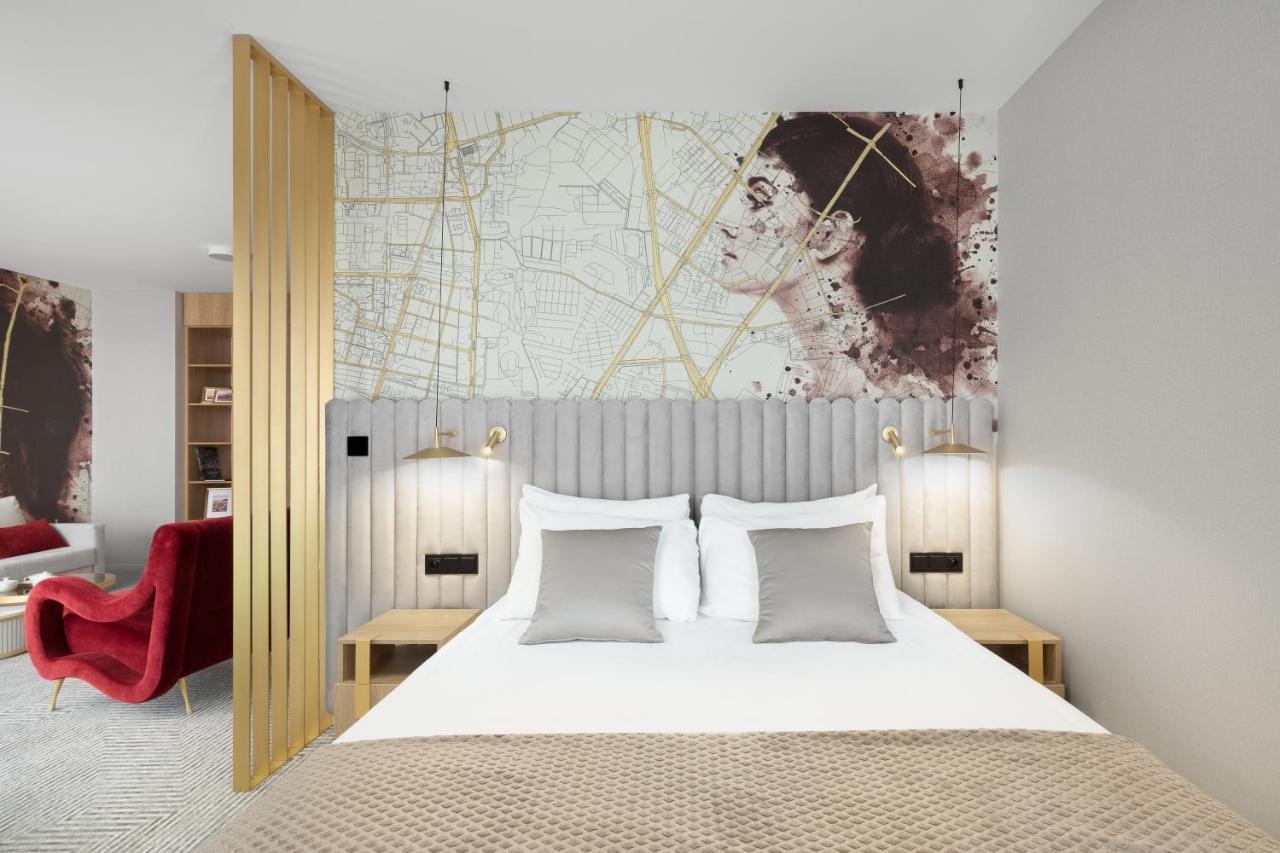 Royal Tulip Warsaw Centre 아파트 호텔 외부 사진 The photo shows a stylish hotel room with a modern aesthetic. It features a bed with a light-colored duvet and several decorative pillows in shades of gray. Behind the bed is a unique headboard with vertical striped design. Above the headboard, there