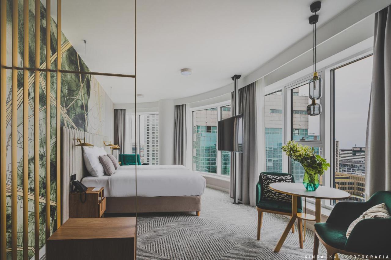 Royal Tulip Warsaw Centre 아파트 호텔 외부 사진 The photo shows a modern hotel room with large windows that let in natural light. The decor features a stylish and contemporary design, including a bed with a white comforter and elegant headboard. There's a wooden nightstand beside the bed and a sma