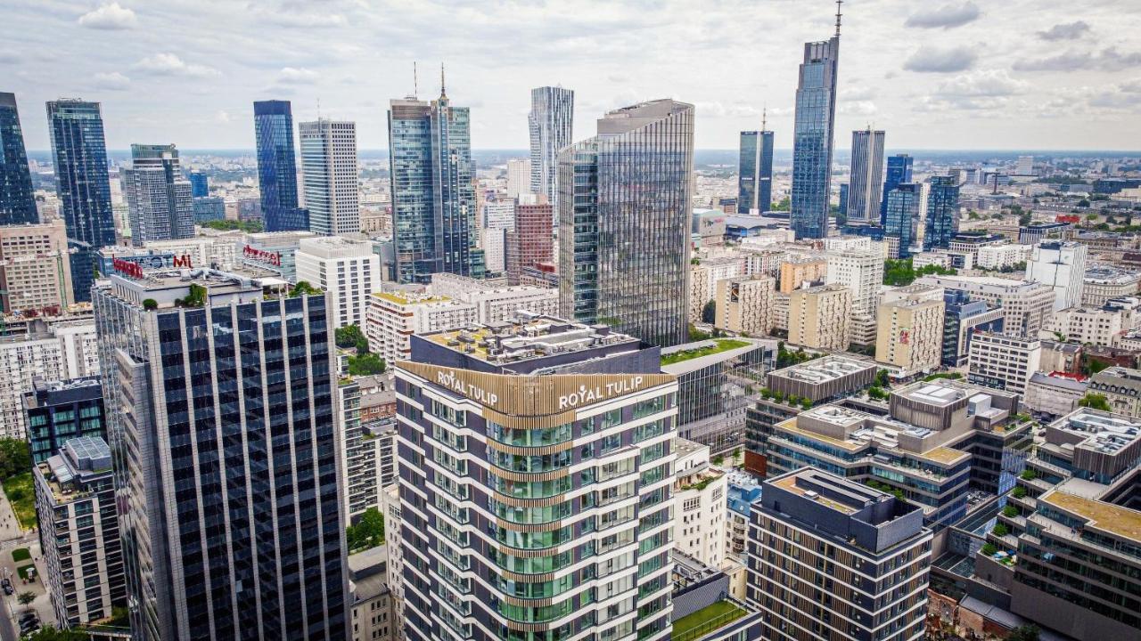 Royal Tulip Warsaw Centre 아파트 호텔 외부 사진 The photo shows a panoramic view of a modern city skyline. In the foreground, there are several tall buildings with contemporary architectural designs. One building prominently features the name "Royal Tulip." The background is filled with numerous s