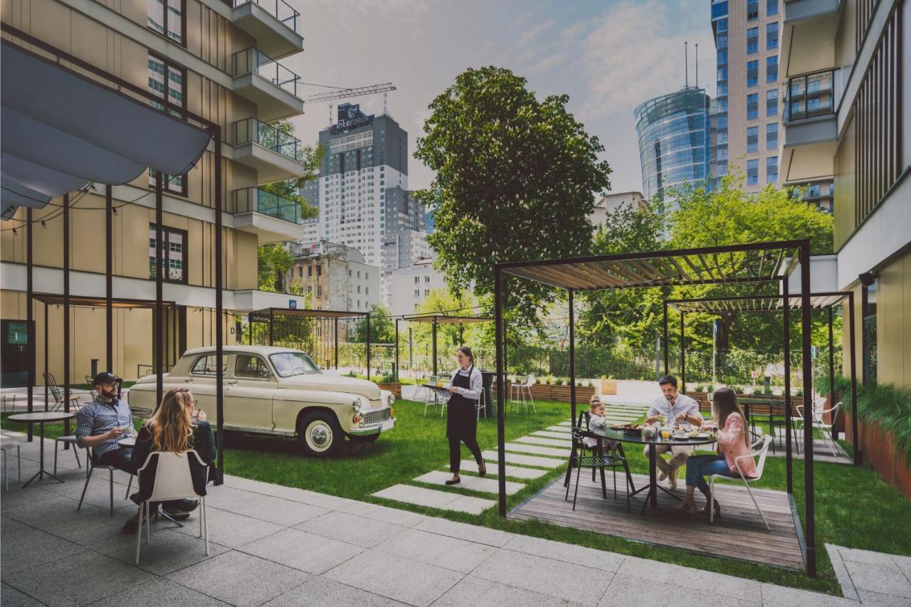 Royal Tulip Warsaw Centre 아파트 호텔 외부 사진 The photo depicts a modern urban setting featuring a green courtyard. In the foreground, there are several people sitting at tables, enjoying a meal or drinks. A couple of individuals are lounging, while others appear to be dining. 

In the backgroun