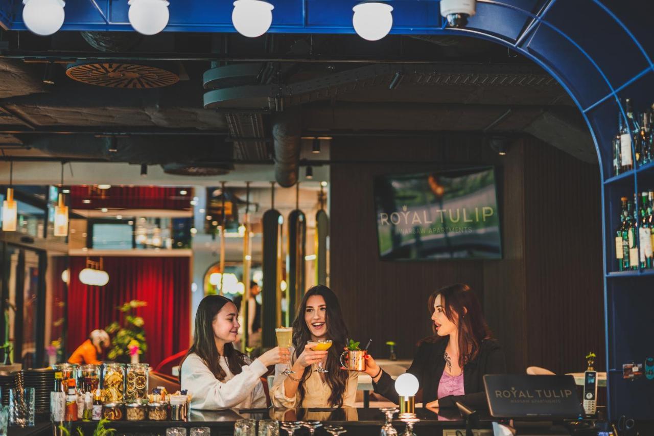 Royal Tulip Warsaw Centre 아파트 호텔 외부 사진 The photo depicts three women gathered around a bar, enjoying drinks. They are smiling and engaged in conversation, exuding a festive and cheerful atmosphere. The bar has a modern design, with colorful decor and lighting. In the background, there is 