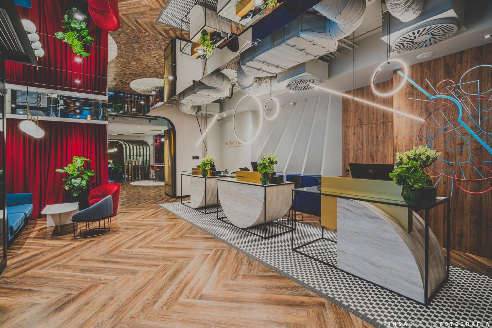 Royal Tulip Warsaw Centre 아파트 호텔 외부 사진 The photo shows a modern hotel lobby with a stylish design. It features a reception area with two main desks, each adorned with plants. The floor has a patterned wooden design, and there are colorful chairs placed around the space. The ceiling is dec