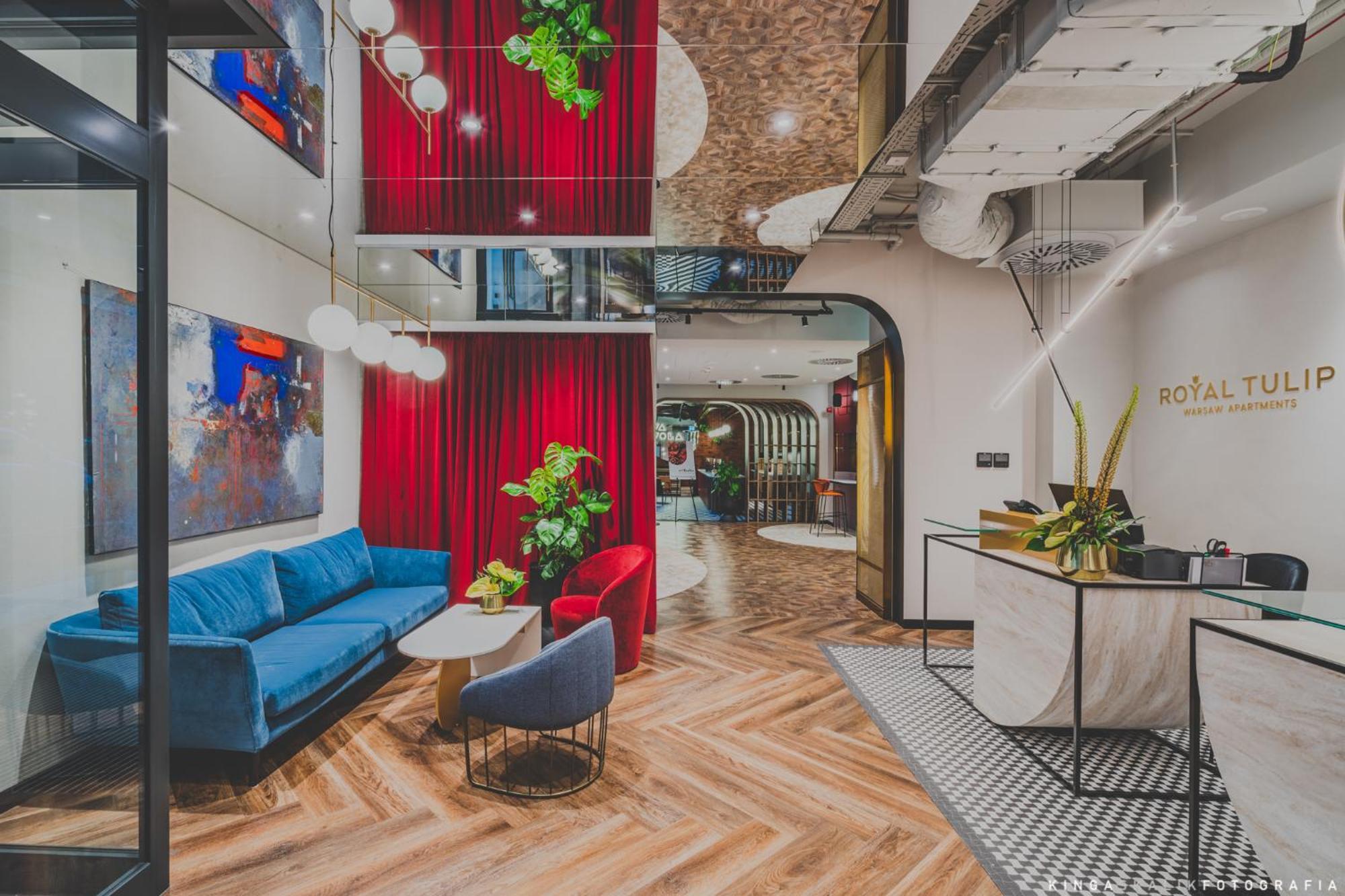 Royal Tulip Warsaw Centre 아파트 호텔 외부 사진 The photo showcases a modern interior space, likely a lobby or lounge area. It features a mix of textures and colors, including a blue couch and red accent chairs. Behind the seating area, there are vibrant red curtains and greenery that adds a touch