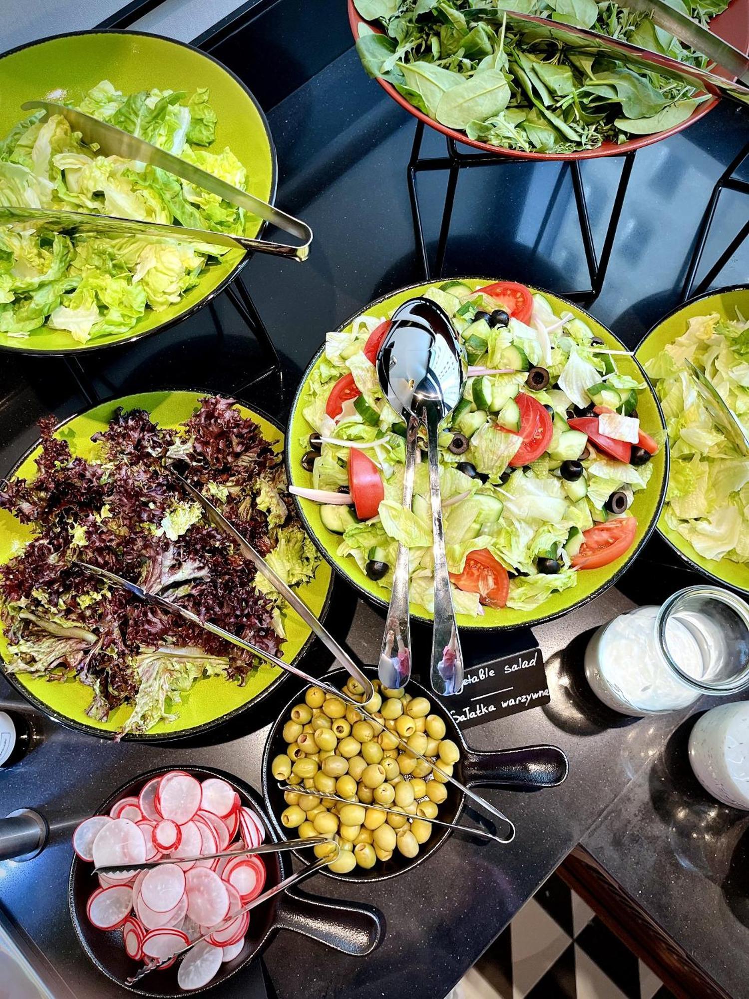 Royal Tulip Warsaw Centre 아파트 호텔 외부 사진 The photo shows a variety of salads displayed in bowls. There are two main salad bowls:

1. **Left Bowl**: Contains dark leafy greens, possibly a mix of different kinds of lettuce or salad greens.
  
2. **Right Bowl**: Features a colorful salad with 
