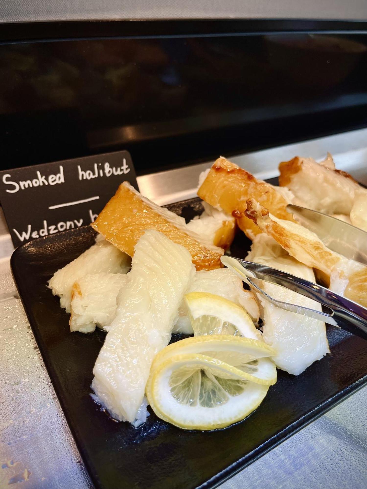 Royal Tulip Warsaw Centre 아파트 호텔 외부 사진 The photo shows a platter of smoked halibut. The fish is presented in white, flaky pieces, some of which have a golden-brown crust. There are slices of lemon on the side, which are often used as a garnish or flavor enhancer. A small sign reads "Smoke