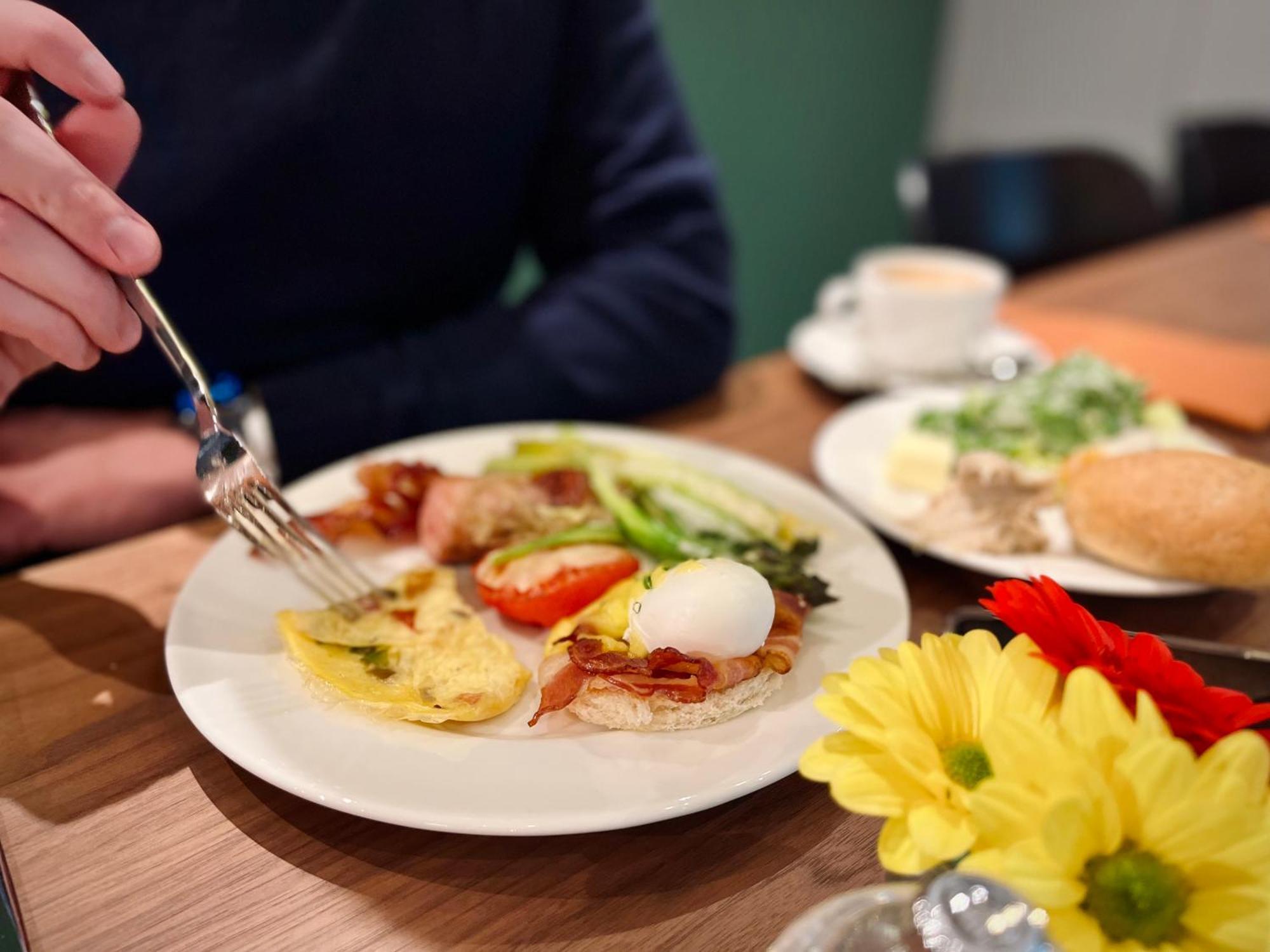 Royal Tulip Warsaw Centre 아파트 호텔 외부 사진 The photo displays a breakfast plate with various items. There's an omelet, a poached egg, bacon, sliced tomatoes, and some greens. A hand is reaching for the food with a fork. Additionally, there are flowers on the table and a small cup of coffee. T