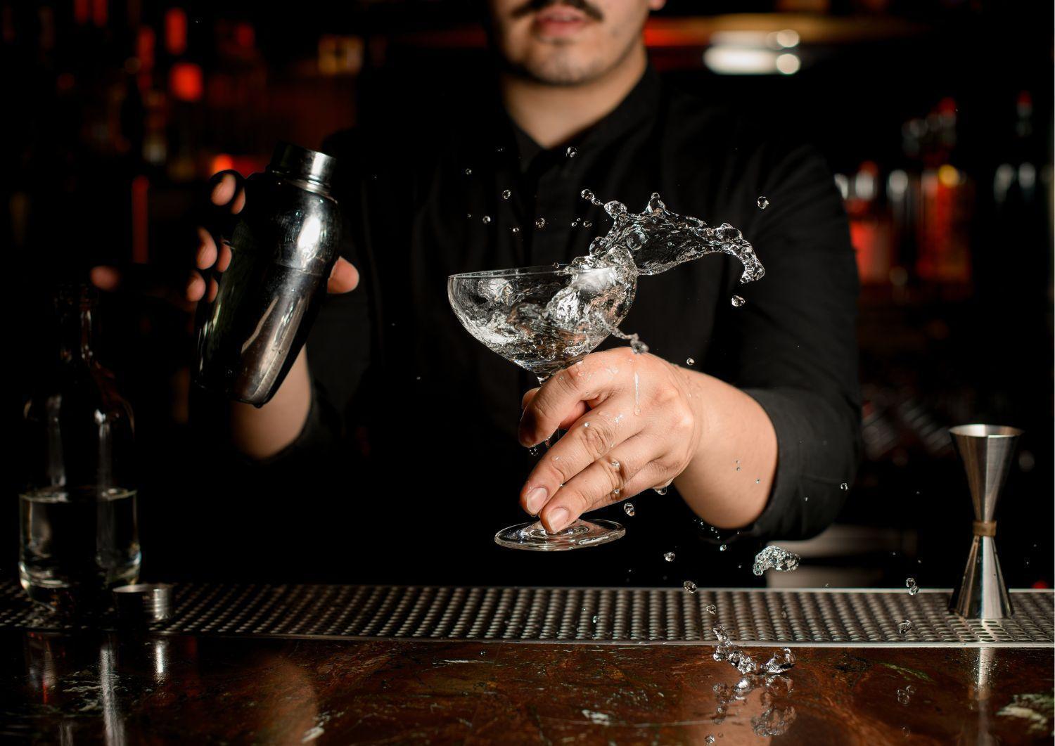 Royal Tulip Warsaw Centre 아파트 호텔 외부 사진 The image shows a bartender in a black shirt shaking a cocktail shaker with one hand. In the other hand, they are holding a glass, likely a cocktail glass, which has liquid splashing out of it. The setting appears to be dimly lit, with blurred lights