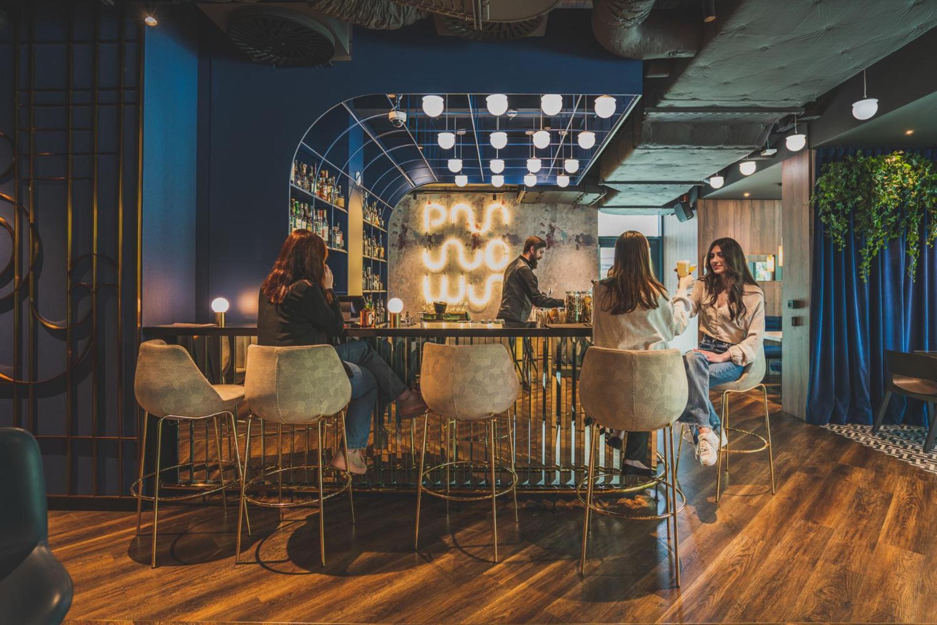 Royal Tulip Warsaw Centre 아파트 호텔 외부 사진 The photo depicts a modern, stylish lounge or bar interior. There are several people sitting at a curved bar with high-back stools, engaged in conversation. The bar area is well-lit with decorative lighting that creates a warm atmosphere. In the back