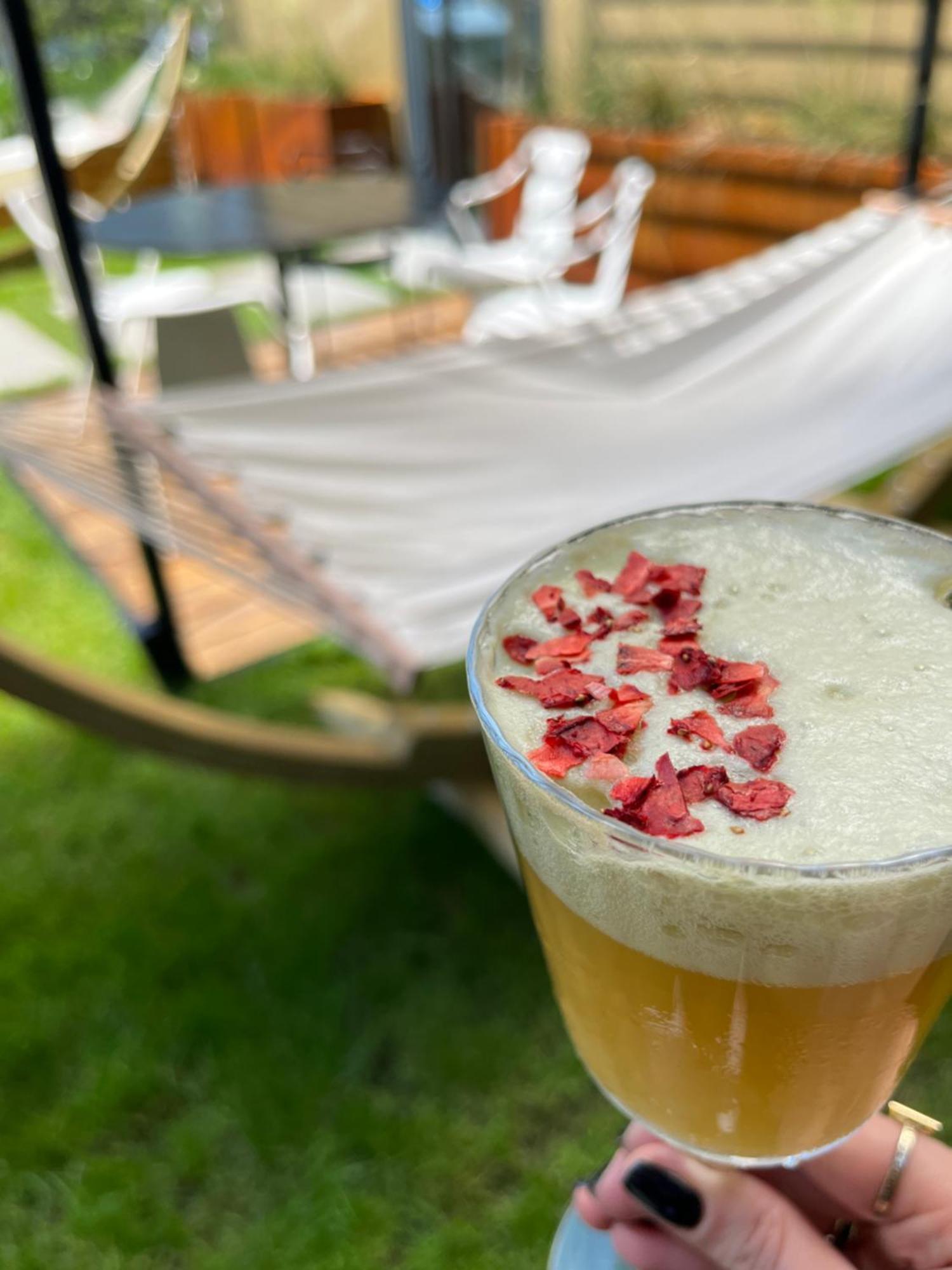 Royal Tulip Warsaw Centre 아파트 호텔 외부 사진 The photo shows a close-up of a drink in a glass, topped with small pieces of red petals or dried flowers. The drink appears to have a frothy white layer above a golden-yellow liquid. In the background, there is a hammock set up on the grass, indicat