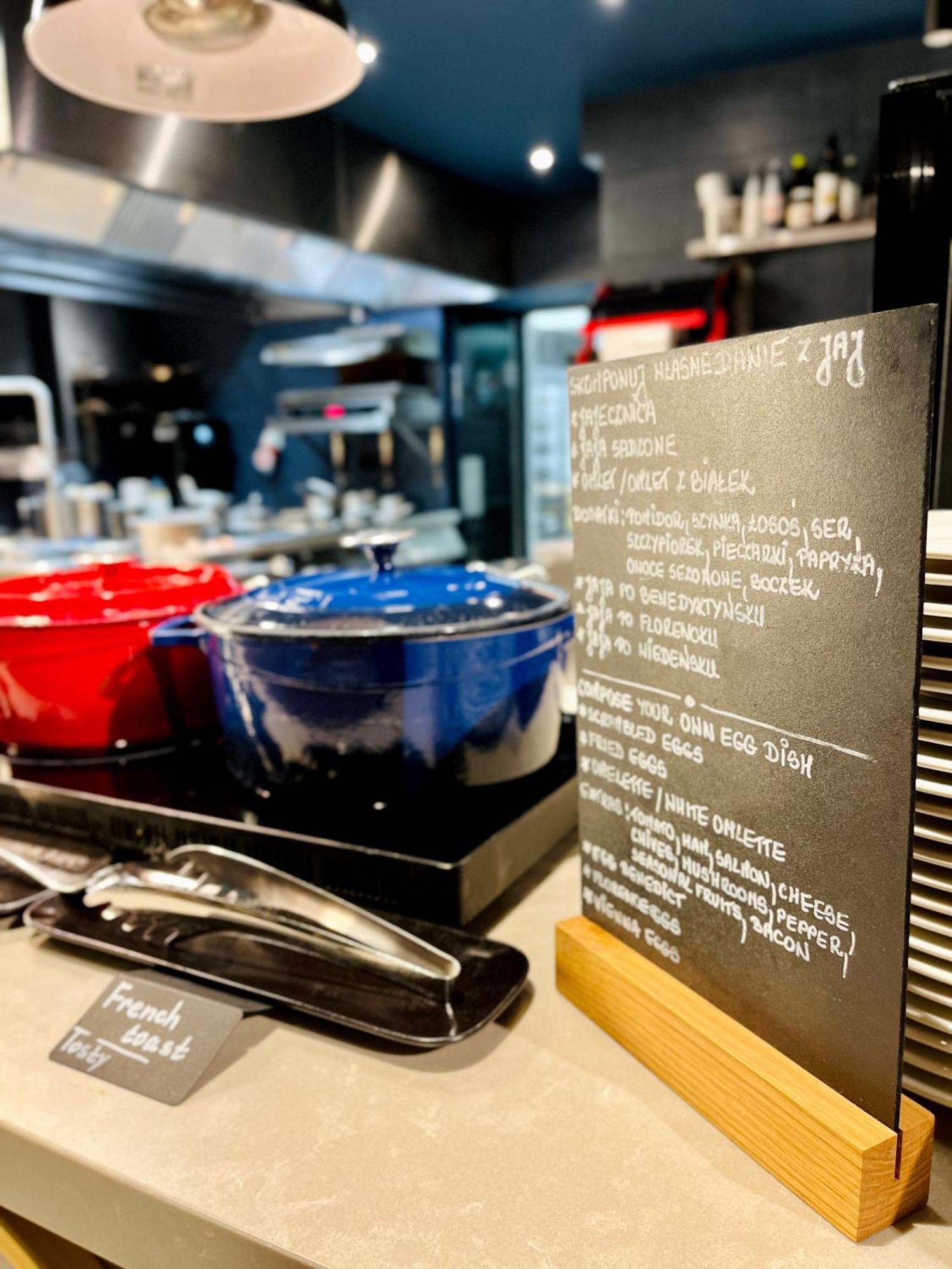 Royal Tulip Warsaw Centre 아파트 호텔 외부 사진 The photo shows a modern kitchen setting with two large pots in the foreground—one red and one blue—placed on a countertop. Next to the pots, there is a menu or chalkboard listing various dishes. The board is decorated with handwritten notes in a lig
