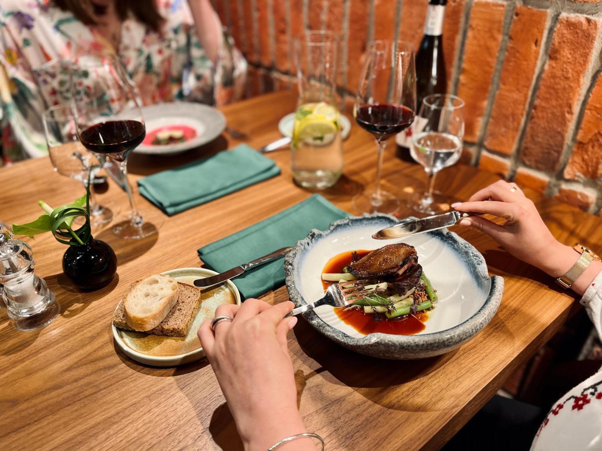 Royal Tulip Warsaw Centre 아파트 호텔 외부 사진 The photo shows a dining scene at a restaurant. There’s a beautifully presented plate of food in the foreground, featuring a piece of meat placed on a bed of greens, garnished with sauce. A person's hand is holding a fork and knife, ready to cut into