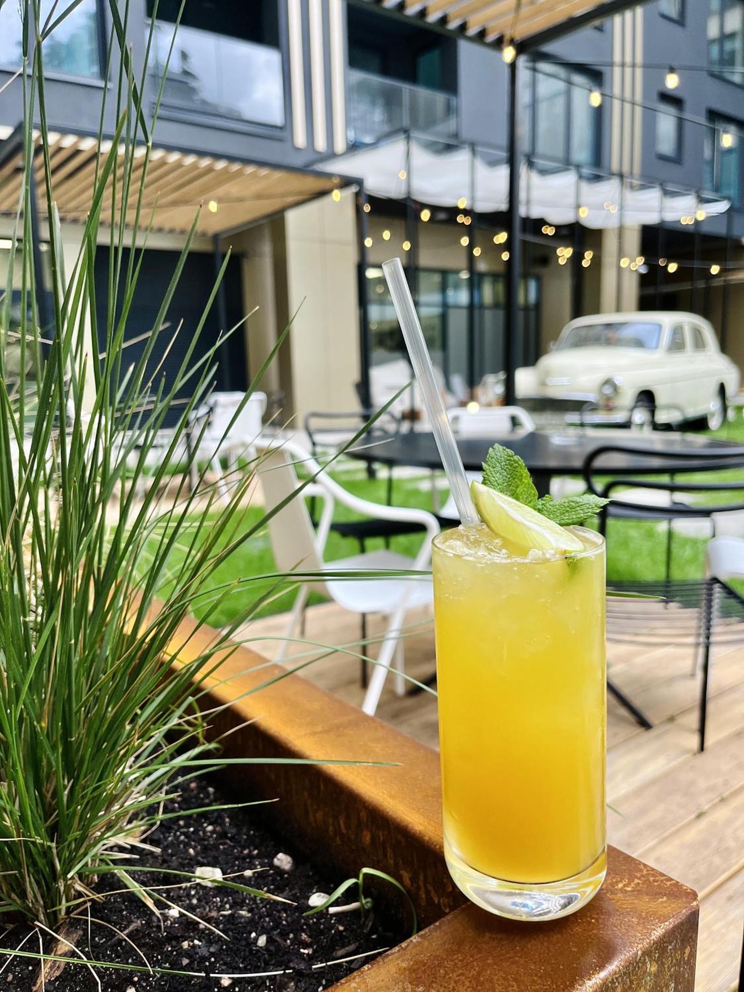 Royal Tulip Warsaw Centre 아파트 호텔 외부 사진 The image shows a refreshing cocktail placed prominently in the foreground, featuring a vibrant yellow color, topped with a slice of lime and a sprig of mint. There is a clear straw inserted into the drink. In the background, there are outdoor seatin