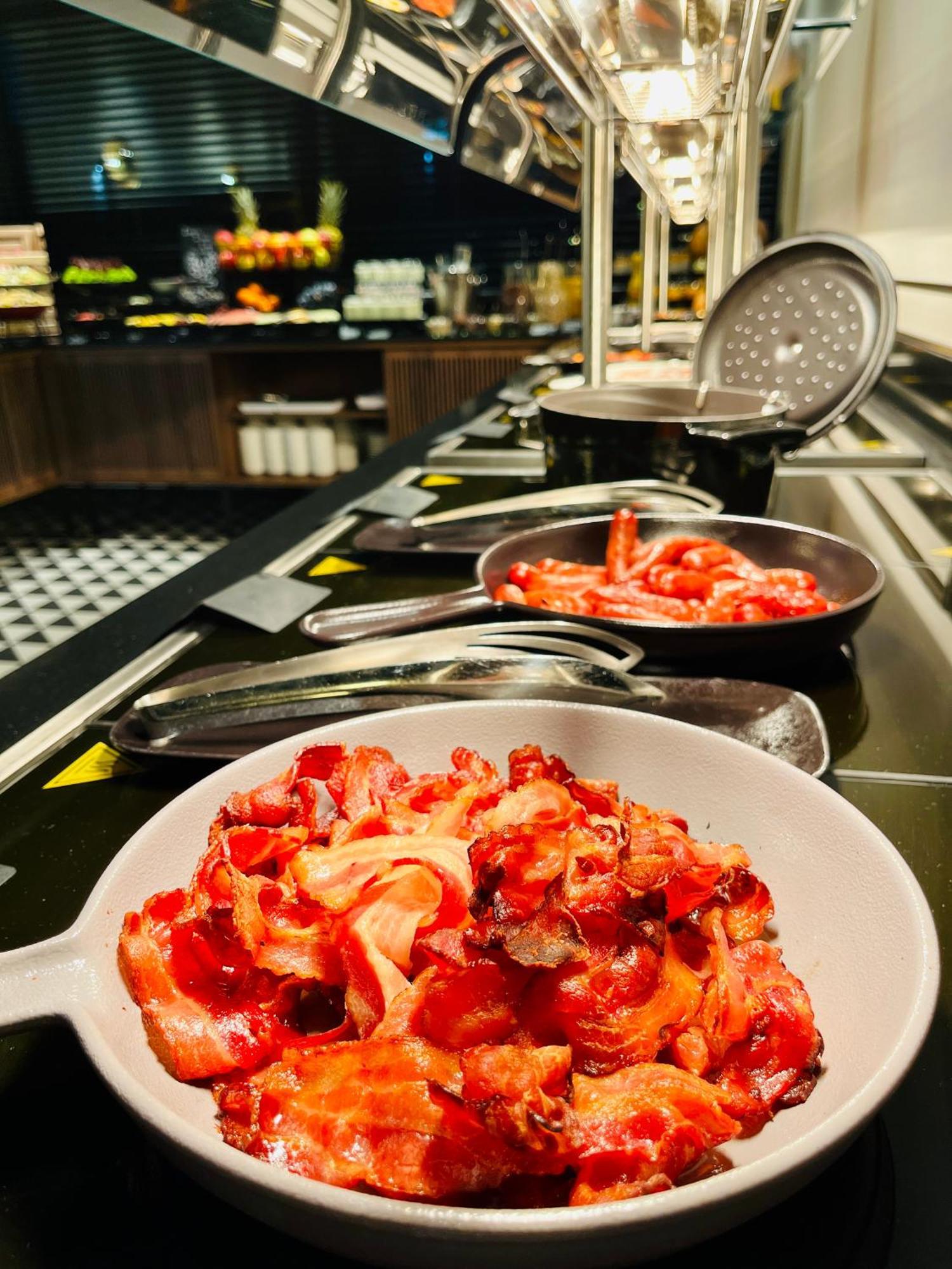 Royal Tulip Warsaw Centre 아파트 호텔 외부 사진 The photo shows a selection of cooked bacon displayed on a buffet or serving station. There are several metal pans with more bacon strips, and one white bowl filled with an ample amount of bacon pieces, all appearing crispy and well-prepared. The set