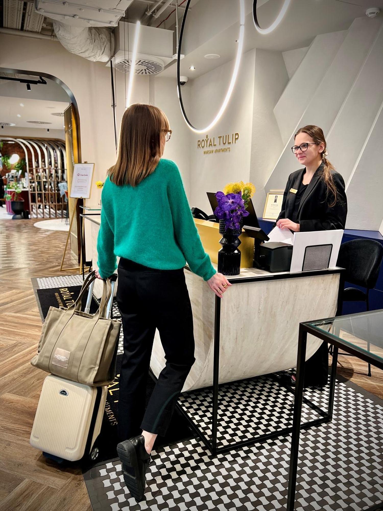 Royal Tulip Warsaw Centre 아파트 호텔 외부 사진 The image shows a hotel reception area. A woman wearing a green sweater and black pants is standing at the reception desk, engaging with a staff member. The staff member, who is smiling and wearing a name tag, is standing behind the desk. There's a b