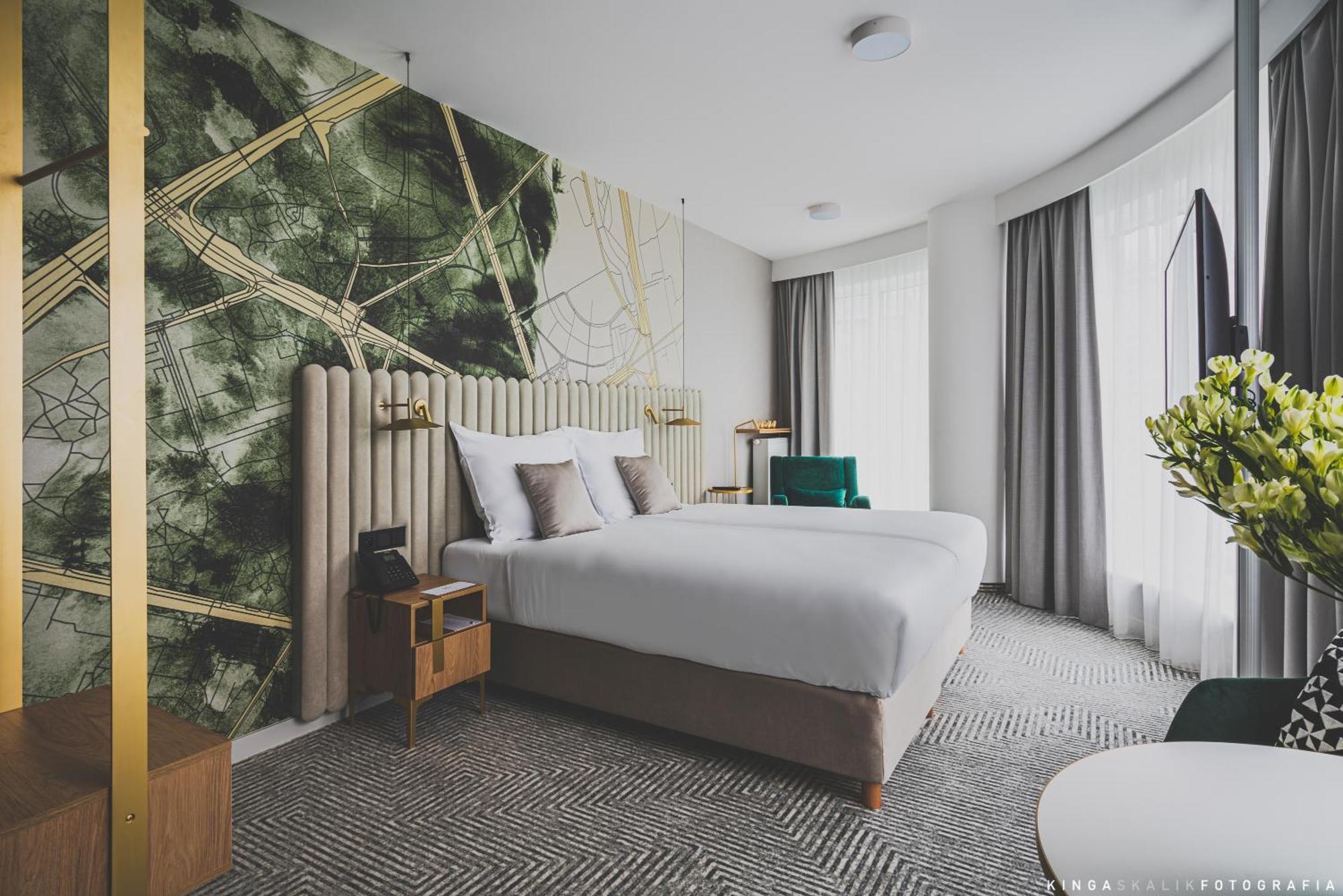 Royal Tulip Warsaw Centre 아파트 호텔 객실 사진 The photo shows a modern hotel room featuring two beds with white bedding. The headboard is upholstered and elegantly designed. Behind the beds, there's a striking wall design with abstract green and gold patterns. The room has large windows with lig